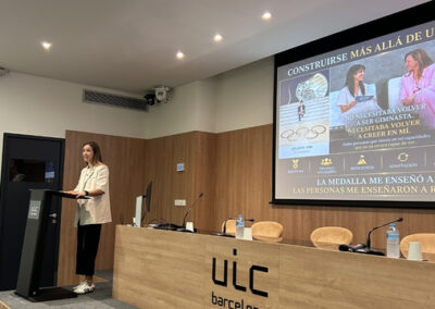 Marta Perarnau interviene desde el atril en una sala de conferencias de UIC Barcelona durante una jornada sobre salud mental y deporte. Tras ella se proyecta una presentación centrada en la resiliencia, la superación personal y el bienestar emocional en el ámbito deportivo, mientras varios asientos y micrófonos ocupan la mesa del auditorio.