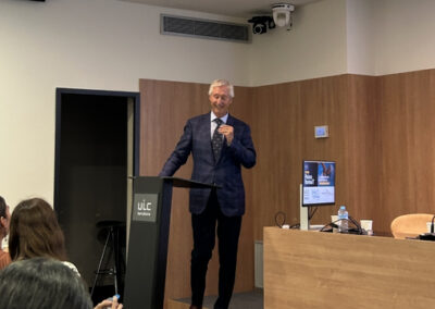 Javier Mirallas, presidente del Patronato de la Fundación Salud y Persona, interviene desde el atril durante una jornada sobre salud mental y deporte celebrada en UIC Barcelona. La imagen muestra al ponente dirigiéndose al público en una sala de conferencias equipada con pantalla de proyección, mesa presidencial y material audiovisual.